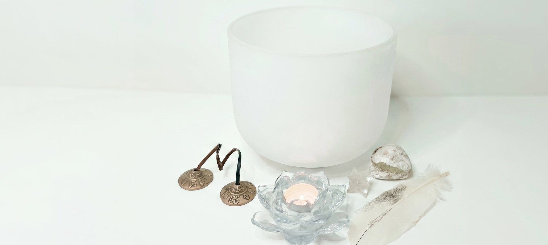 A frosted white singing bowl candle holder with a small tealight candle, surrounded by white crystals, a feather, and two small metallic coins on a white surface against a white background.