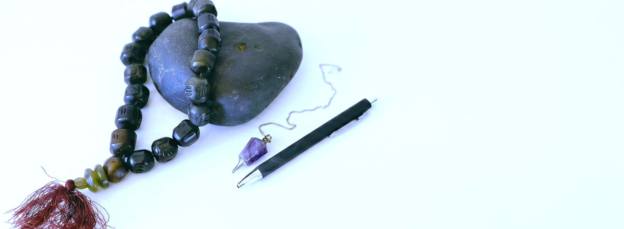 A beaded necklace with dark brown and black beads, a reddish-brown tassel, and a small silver-toned pendant, displayed next to a black pen on a plain white background.